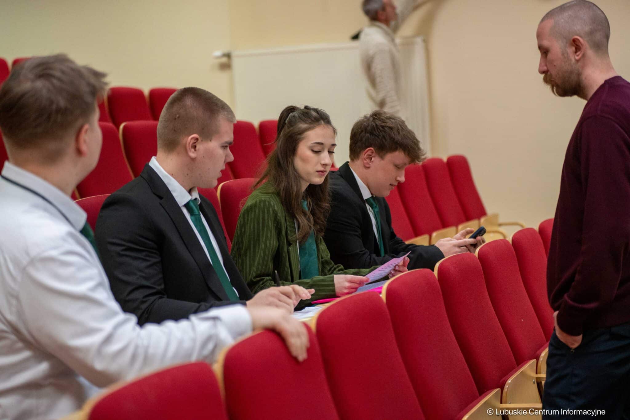 Group of young adults in a lecture hall with red seats; several in suits review papers while a man stands nearby.
