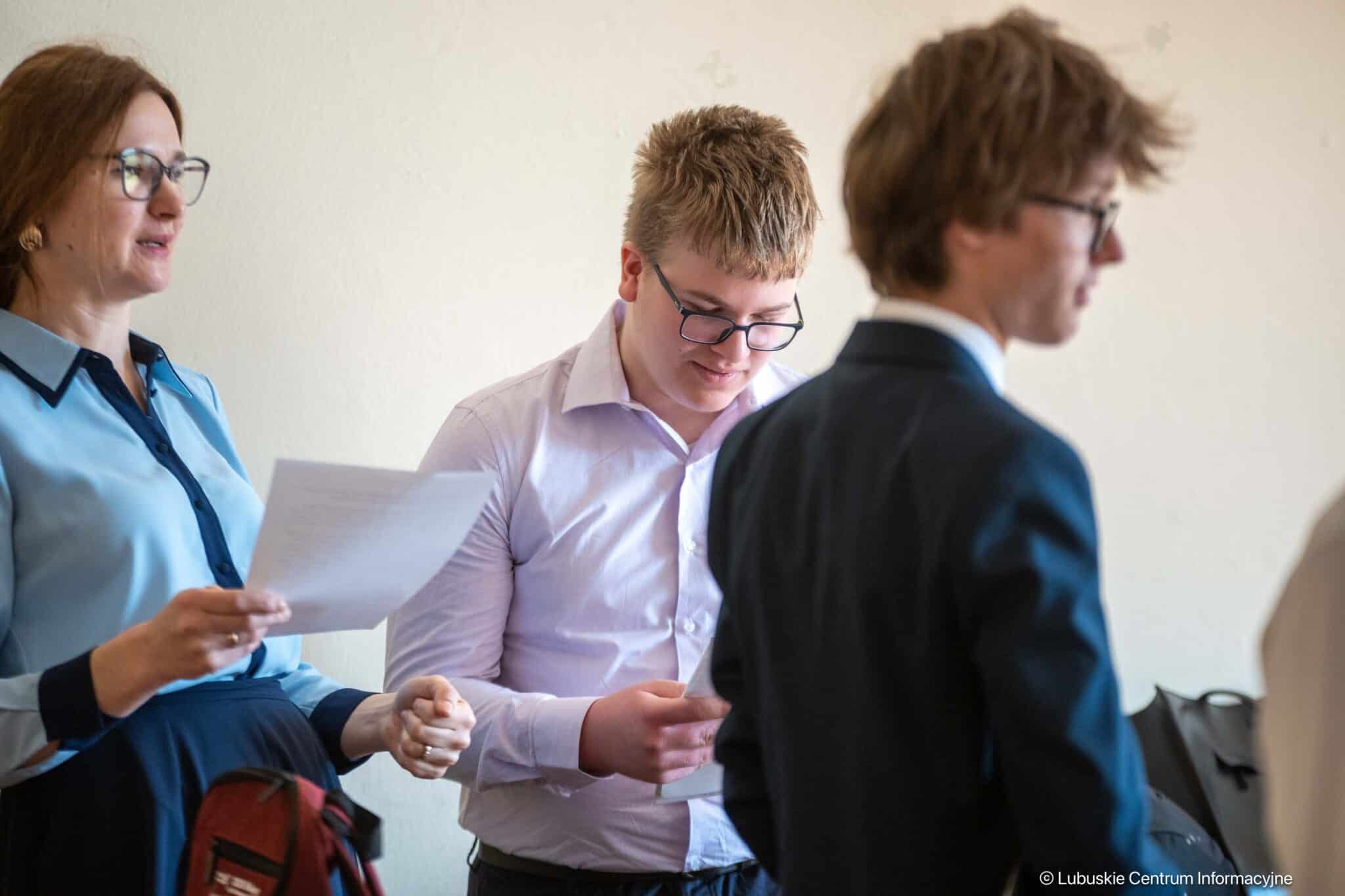 Woman in a blue blouse with glasses holds a sheet of paper as a student in a light shirt reads another document, with a man in a suit nearby (blurry).