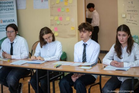 Four students sit at a classroom table with papers and pens, looking serious and focused on their work.