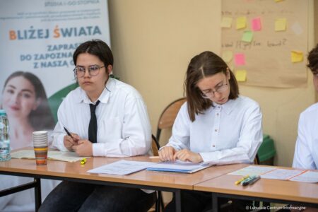 Two students in white shirts sit at a desk, taking notes, with papers, pens, and a water bottle on the table and a banner in the background.