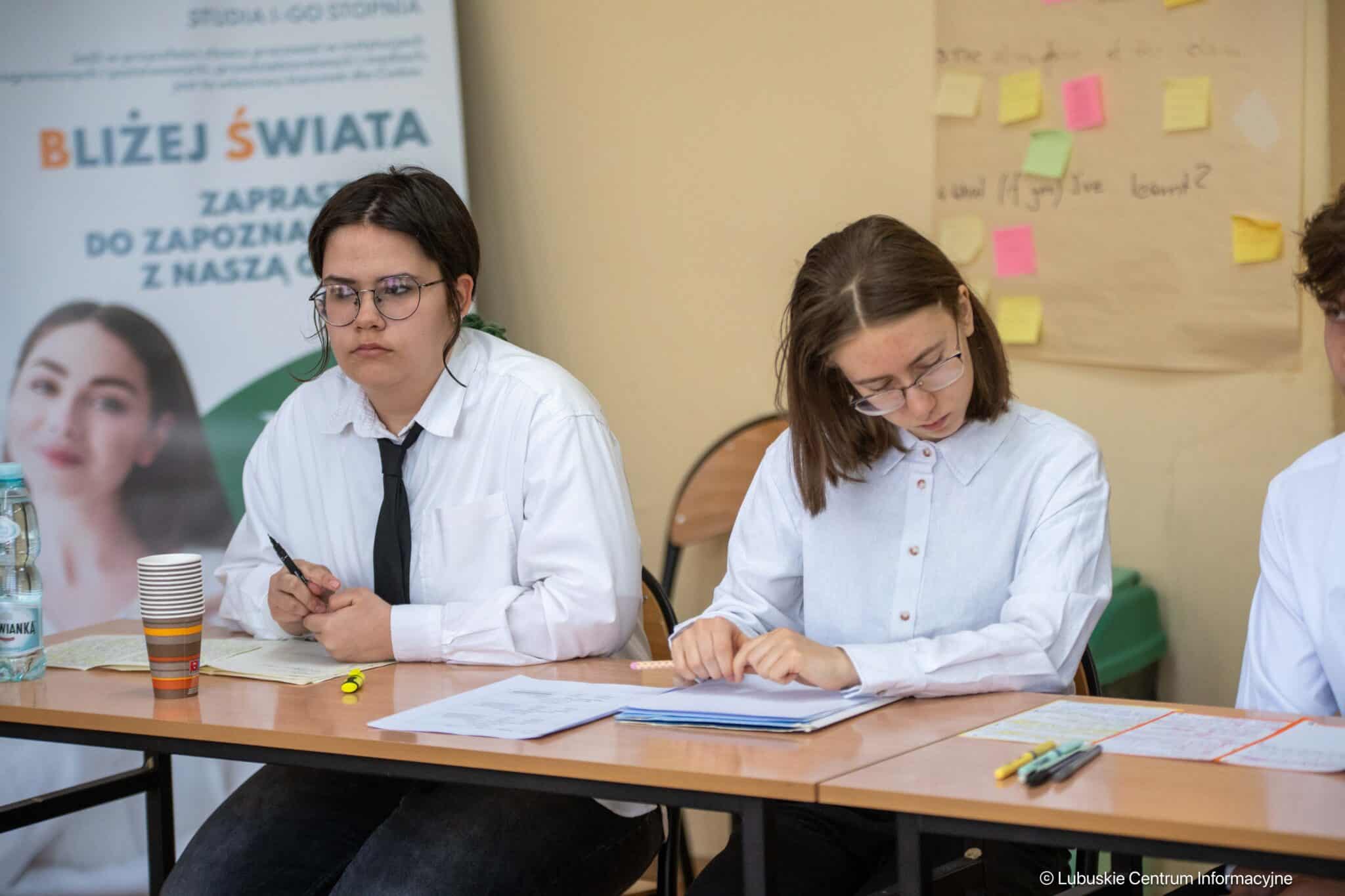 Two students in white shirts sit at a desk, taking notes, with papers, pens, and a water bottle on the table and a banner in the background.