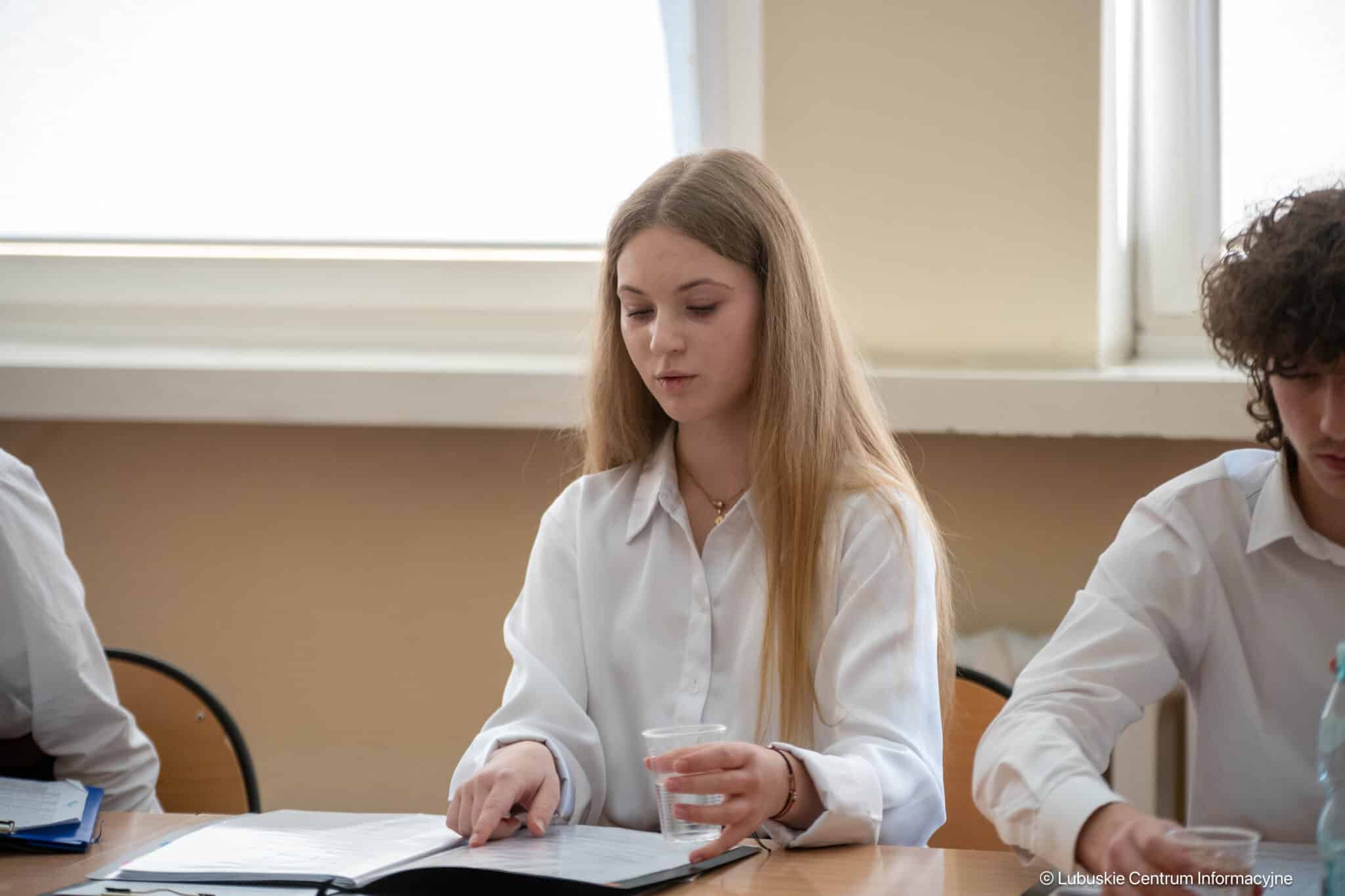 Woman in a white shirt sits at a table, studying papers with a cup in her hand.