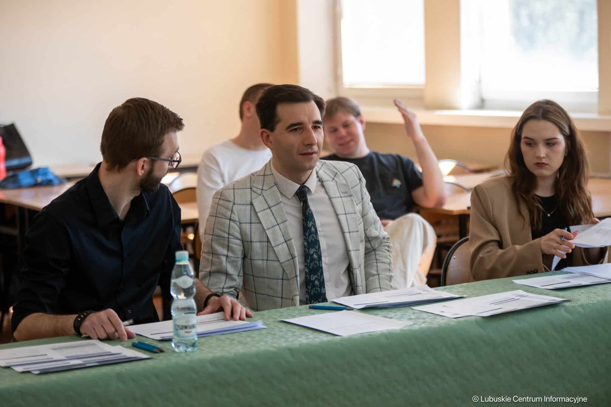 Group of people seated at a classroom table with papers; man in a light plaid blazer center, woman on the right reviewing documents, others in the background.