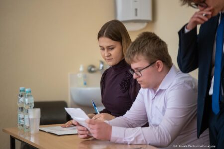 Two people sit at a table reviewing documents, with water bottles and cups nearby as they read and write notes.