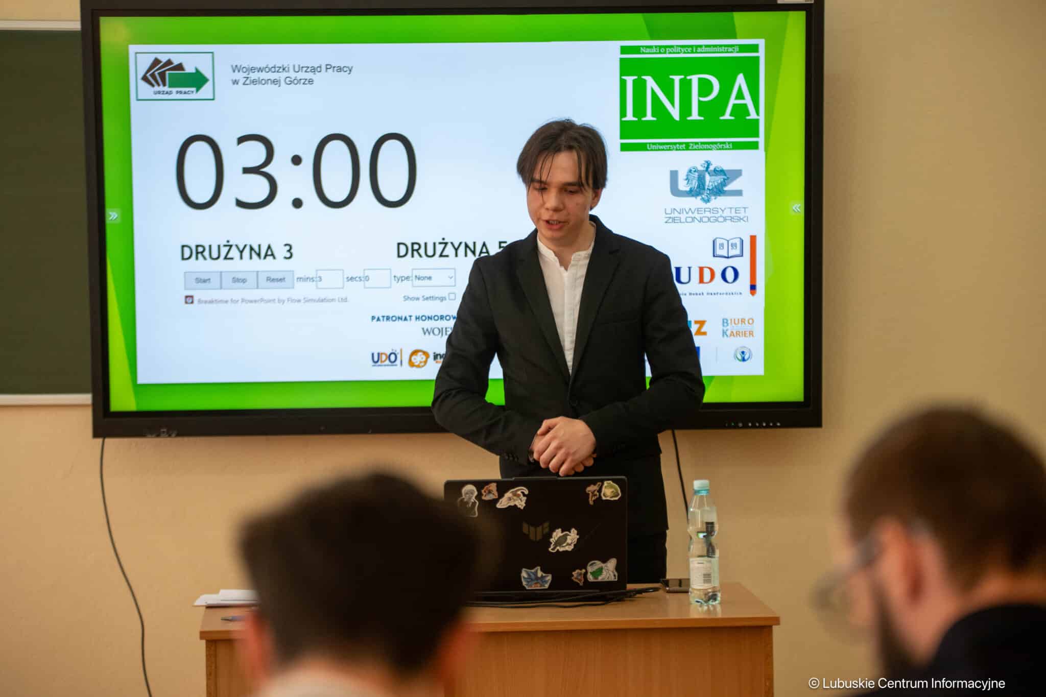 Presenter in a black suit stands at a desk while a large screen shows a 03:00 countdown and logos in the background.