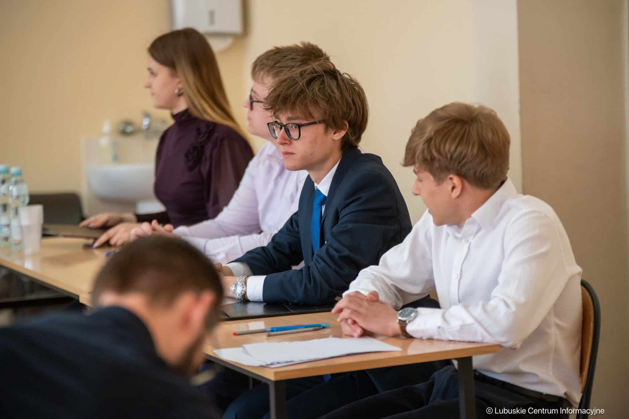 Group of young adults in business attire seated at a conference table, with papers and pens in front, looking attentive.