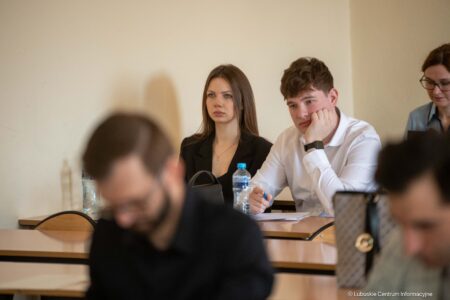 Group of students in a classroom; a young woman in a black blazer sits focused while a young man in a white shirt rests his chin on his hand beside her.