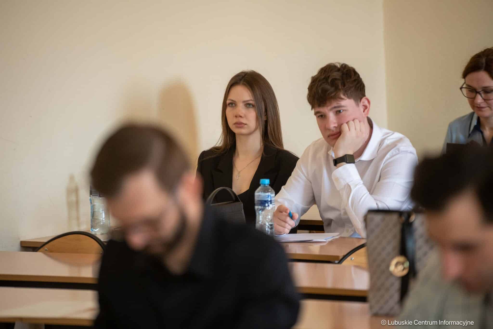 Group of students in a classroom; a young woman in a black blazer sits focused while a young man in a white shirt rests his chin on his hand beside her.