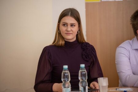 Young woman in a dark purple blouse with a ruffled shoulder sits at a table, with two water bottles and plastic cups in front of her.