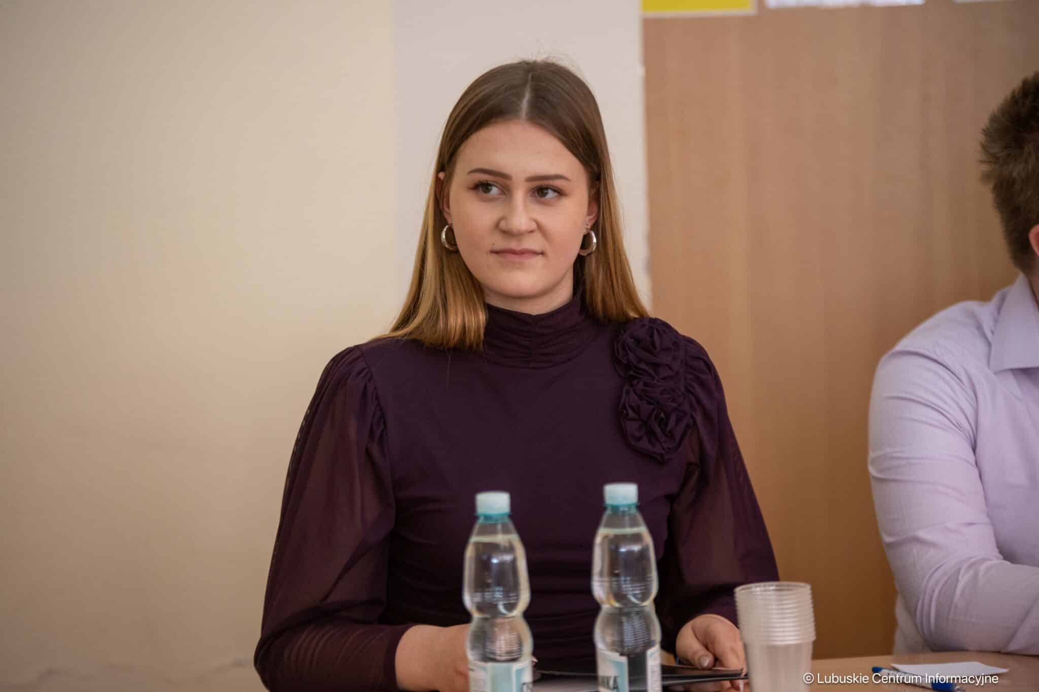 Young woman in a dark purple blouse with a ruffled shoulder sits at a table, with two water bottles and plastic cups in front of her.