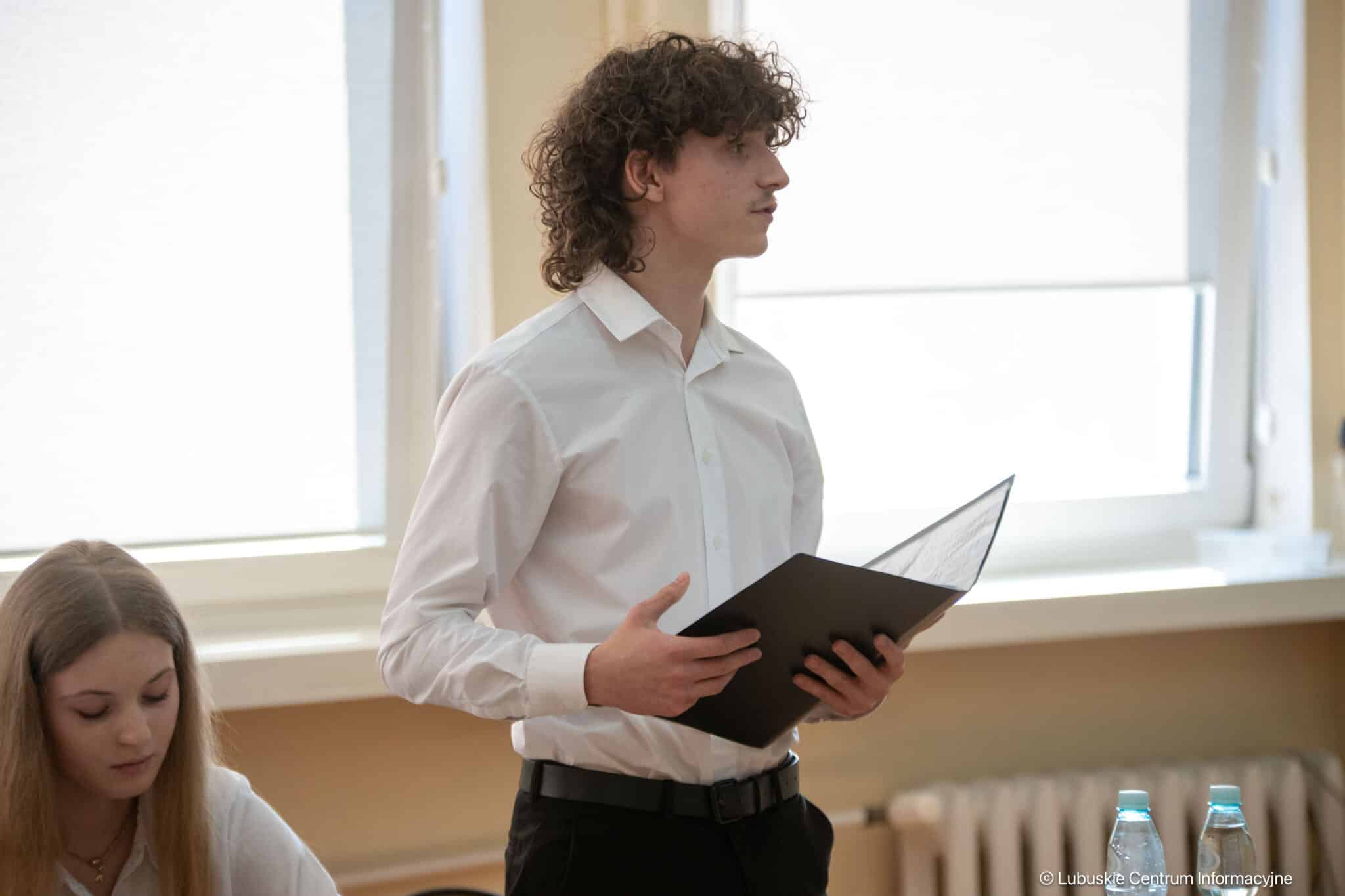 Young man in a white shirt stands and speaks in a classroom, holding a black folder/binder.