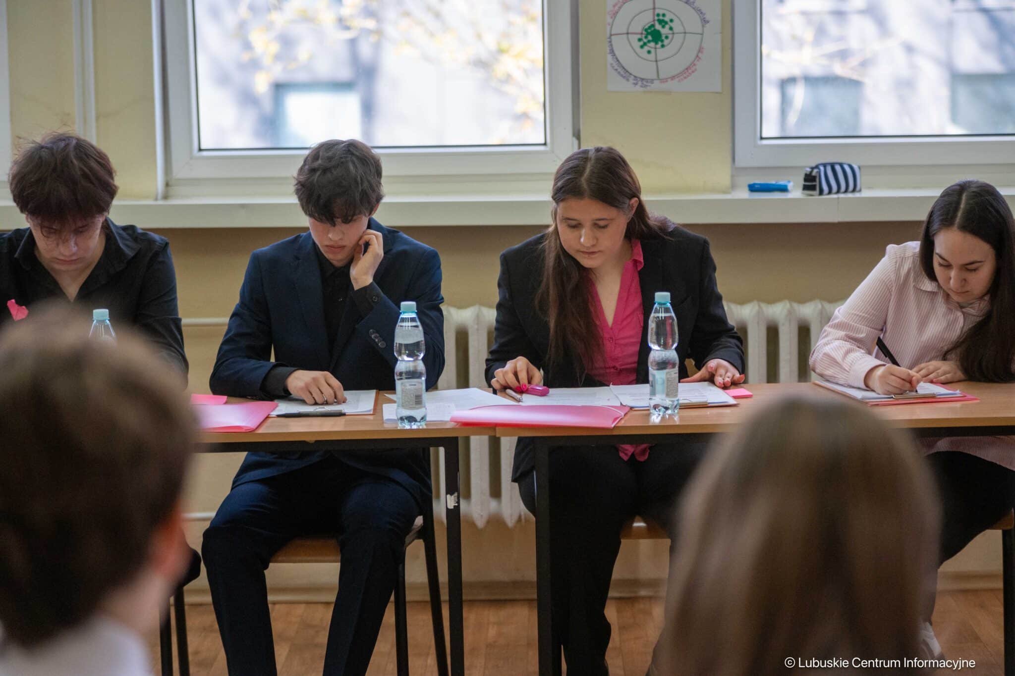 Four students sit at a long table in a classroom, writing on papers with pink folders and bottled water in front of them.