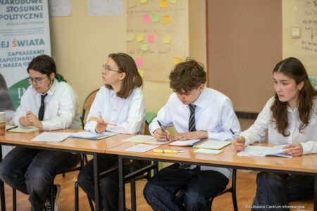 Four students in white shirts sit at a long table, writing notes in a classroom setting.