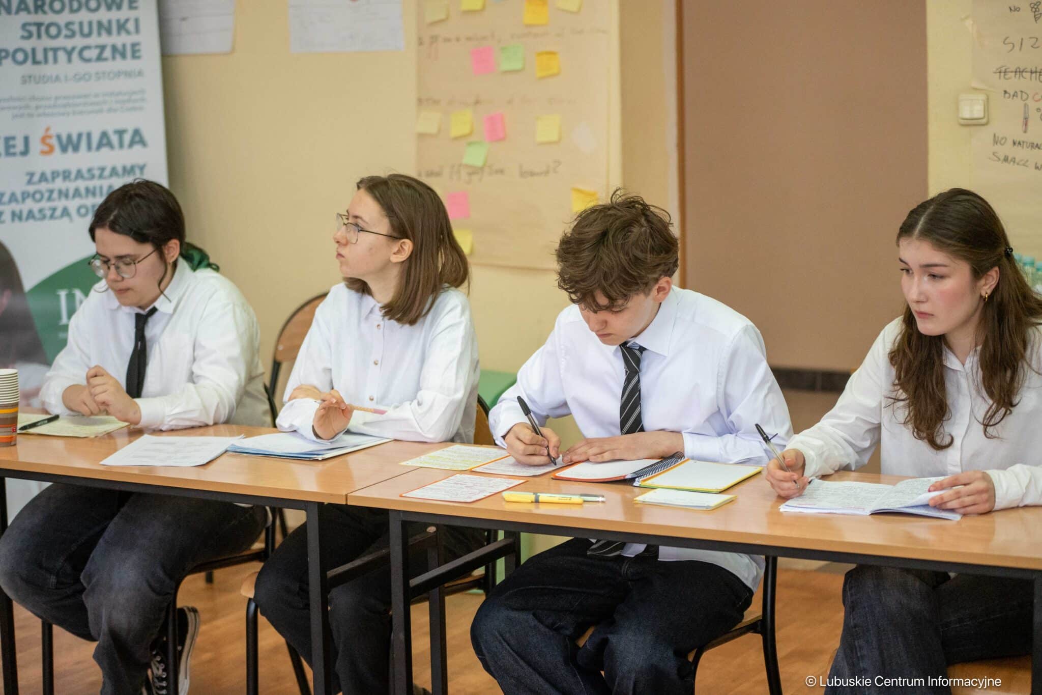 Four students in white shirts sit at a long table, writing notes in a classroom setting.