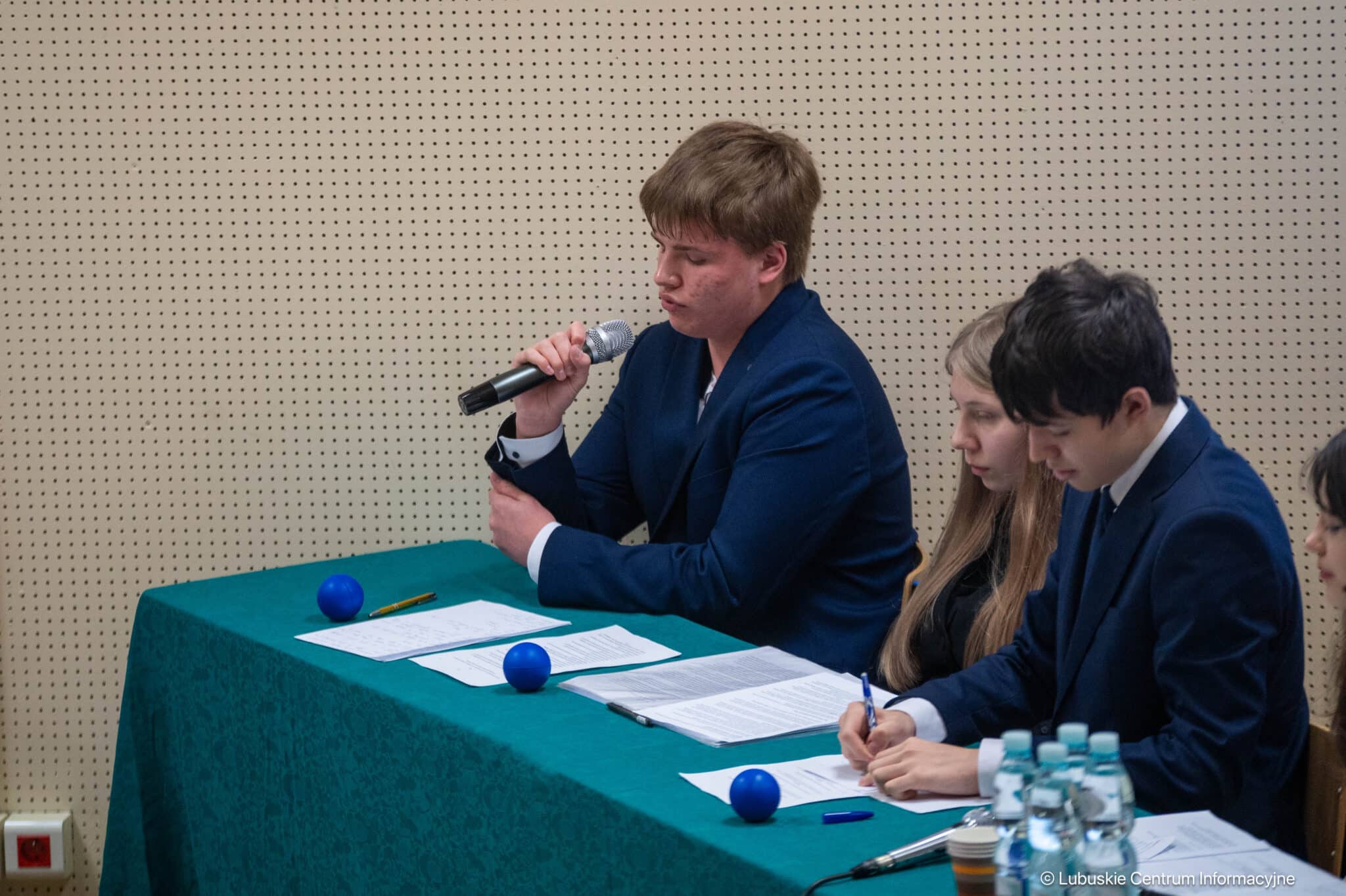 Young man in a blue suit speaks into a microphone at a panel table, with teammates listening and taking notes nearby.