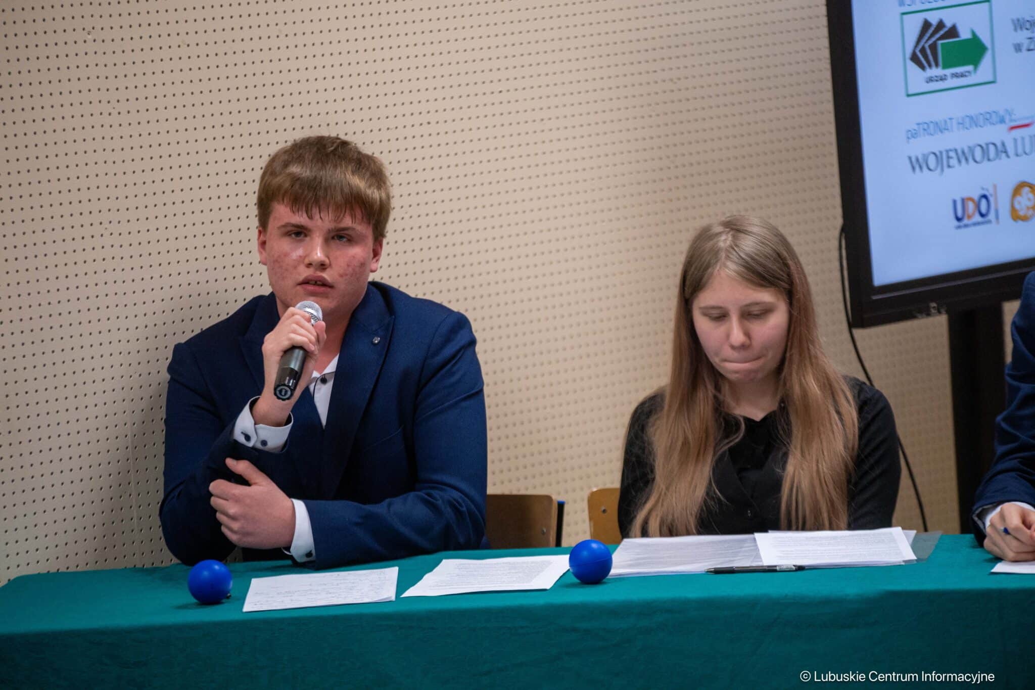 Young man in a blue suit speaks into a handheld microphone at a panel table, with a woman reading papers beside him.