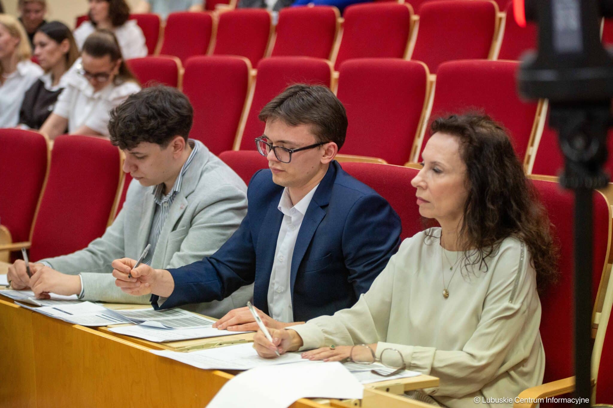 Three adults sit at a wooden desk in a conference room, taking notes with red auditorium seats behind them.