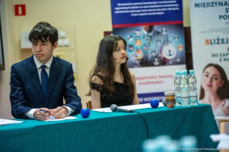 Two young presenters sit at a teal table with papers and a microphone, banners behind them at a conference.