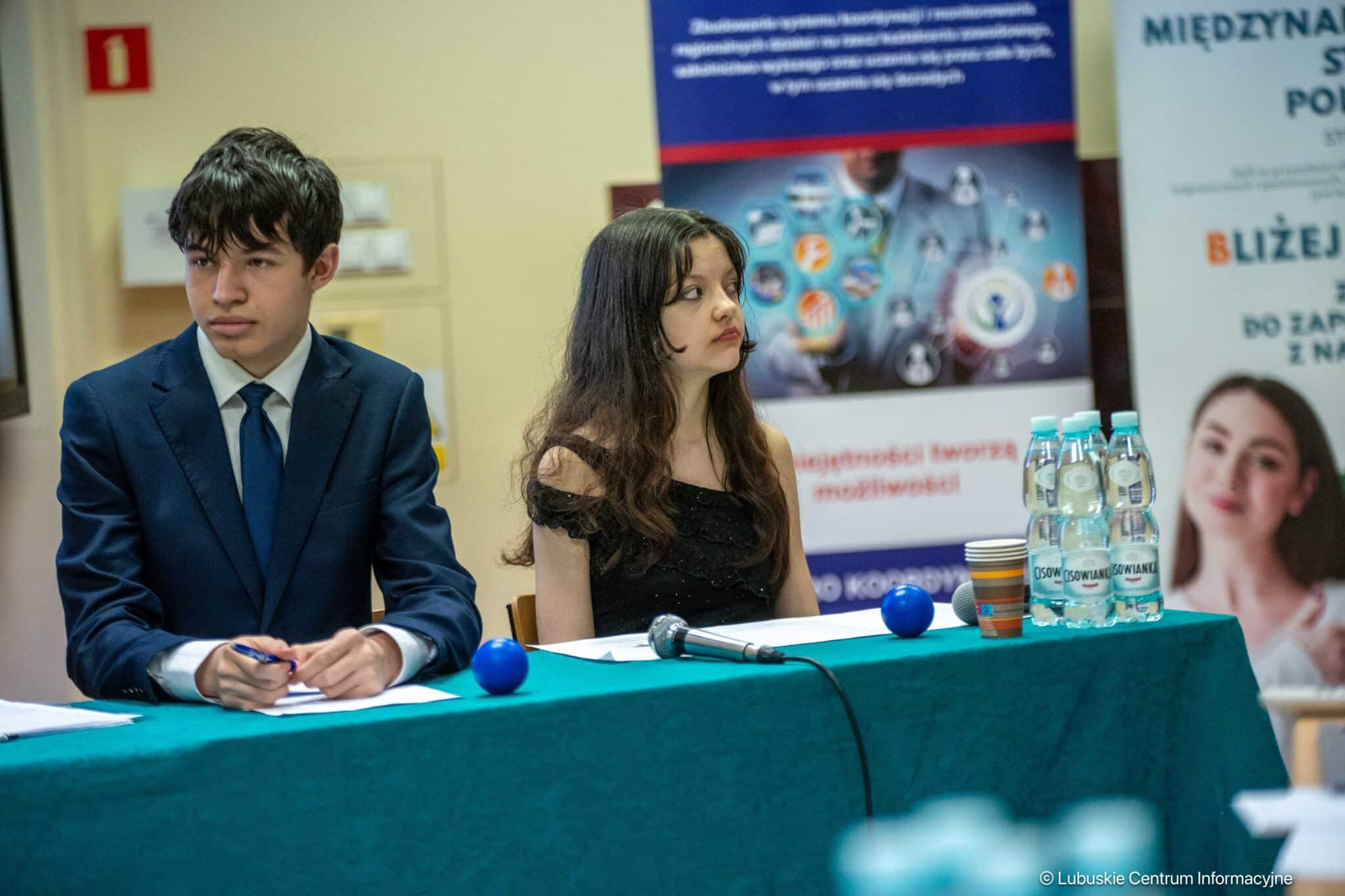 Two young presenters sit at a teal table with papers and a microphone, banners behind them at a conference.