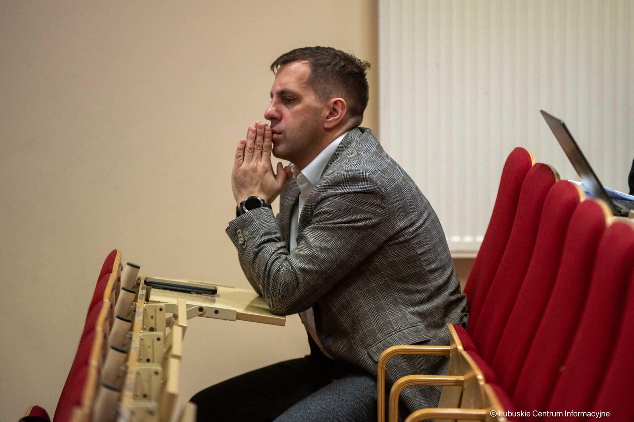 Man in a gray plaid blazer sits in an auditorium, hands pressed together near his lips in contemplation.