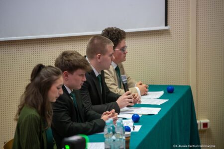 Panel of five people in suits seated at a teal table with papers and a microphone during a discussion session in a conference room.