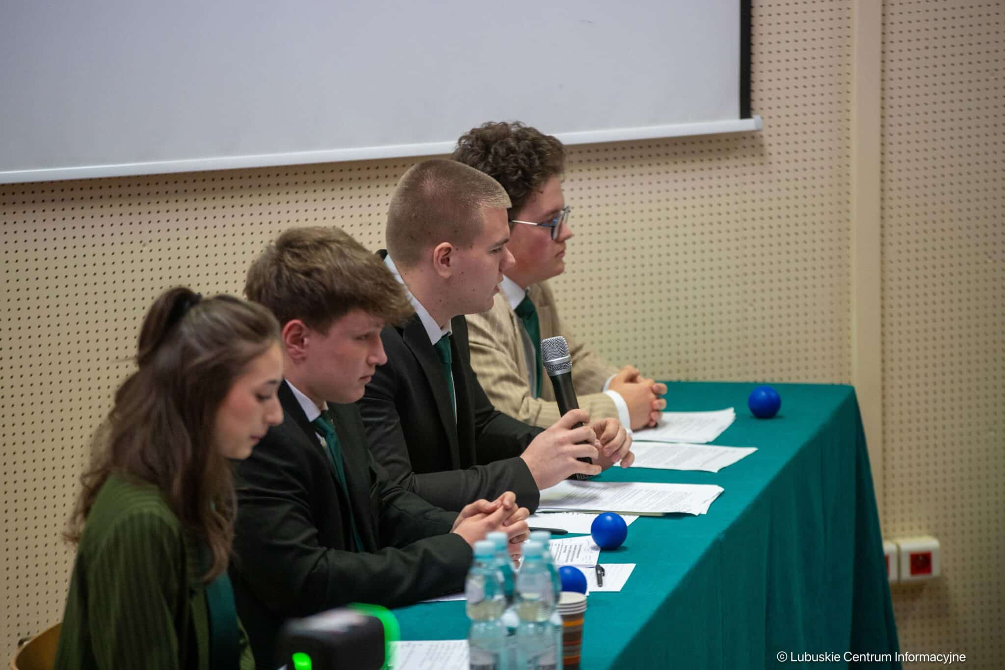Panel of five people in suits seated at a teal table with papers and a microphone during a discussion session in a conference room.