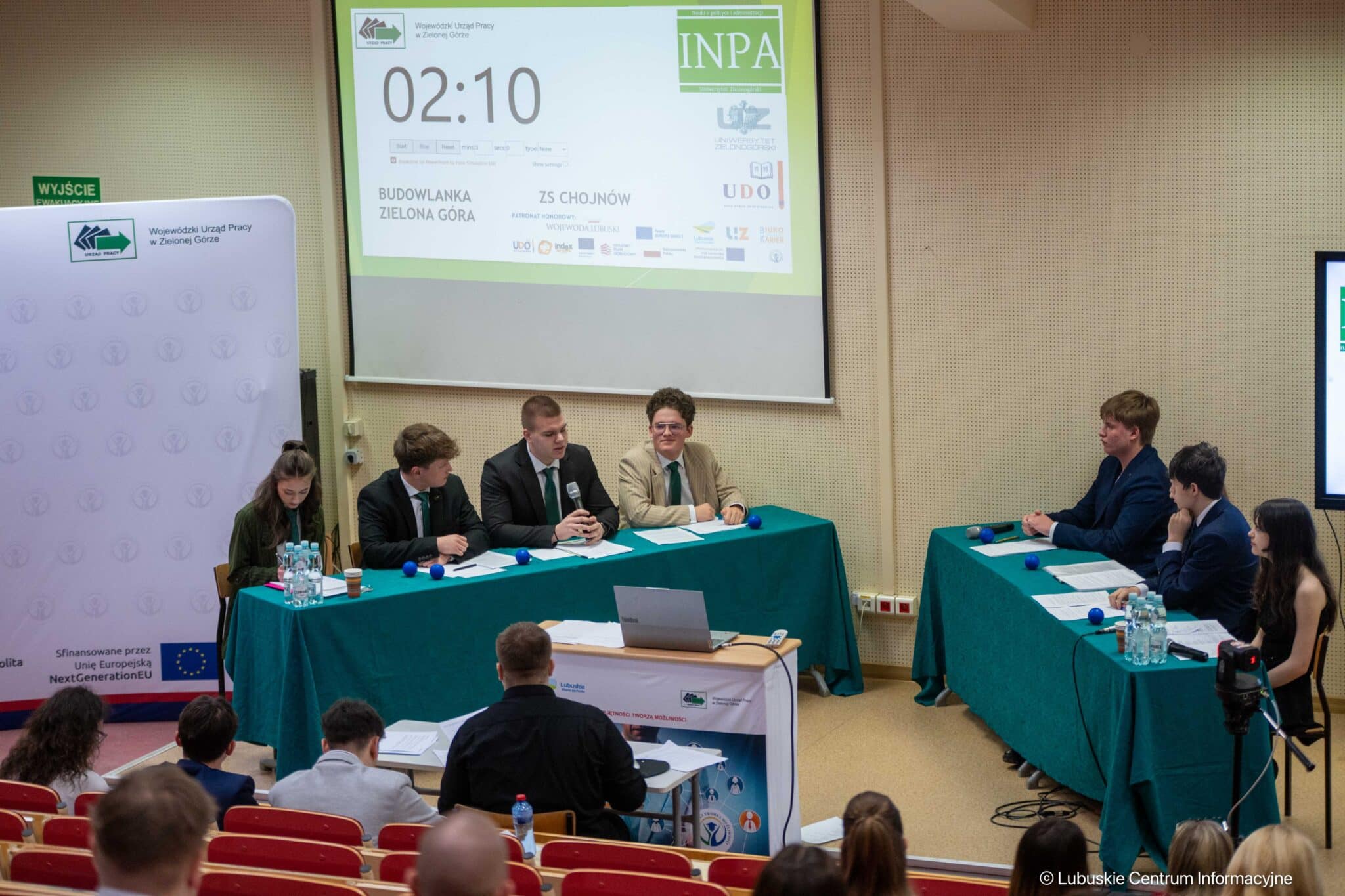 Panel of several young speakers seated at teal tables in a conference room, presenting to an audience with a large projection screen behind them.