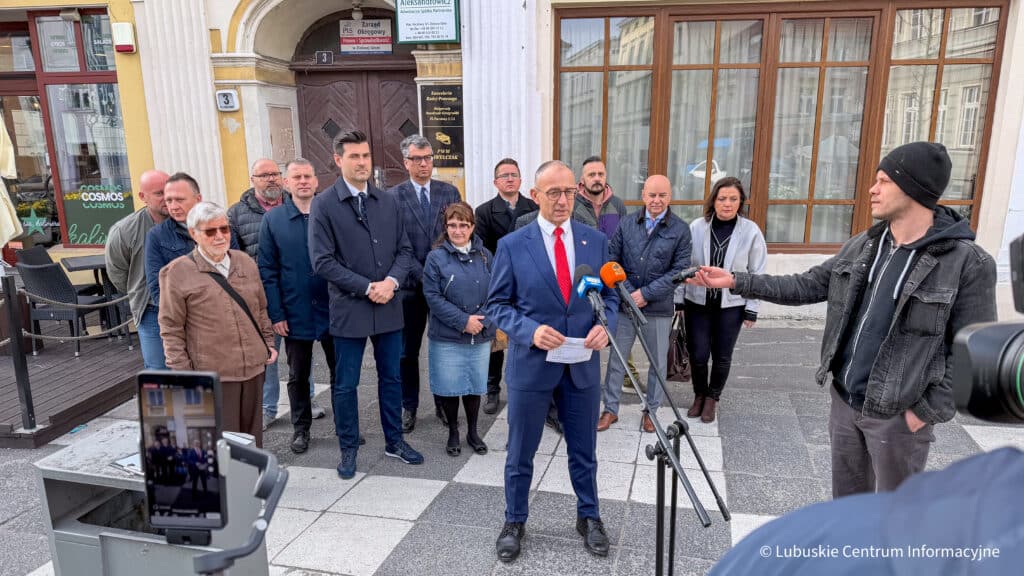 Konferencja przed siedzibą PiS w Zielonej Górze Man in a blue suit delivering a press statement with multiple microphones in front of a doorway surrounded by reporters and onlookers outside a building KO uderza w Mejzę To recydywa i zagrożenie dla mieszkańców