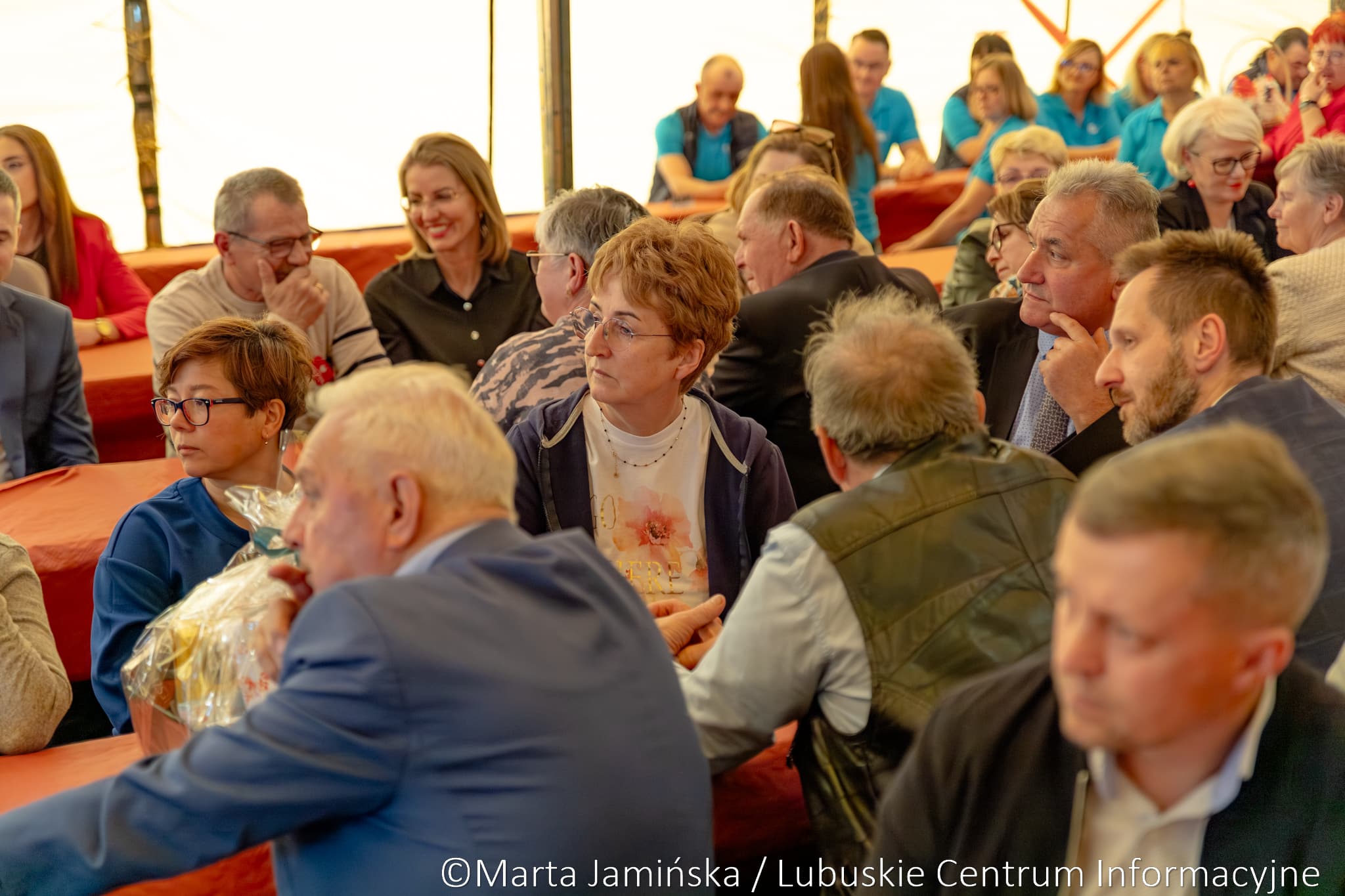 Group of adults seated at orange-covered tables in a bright tent, chatting and listening at a community event.