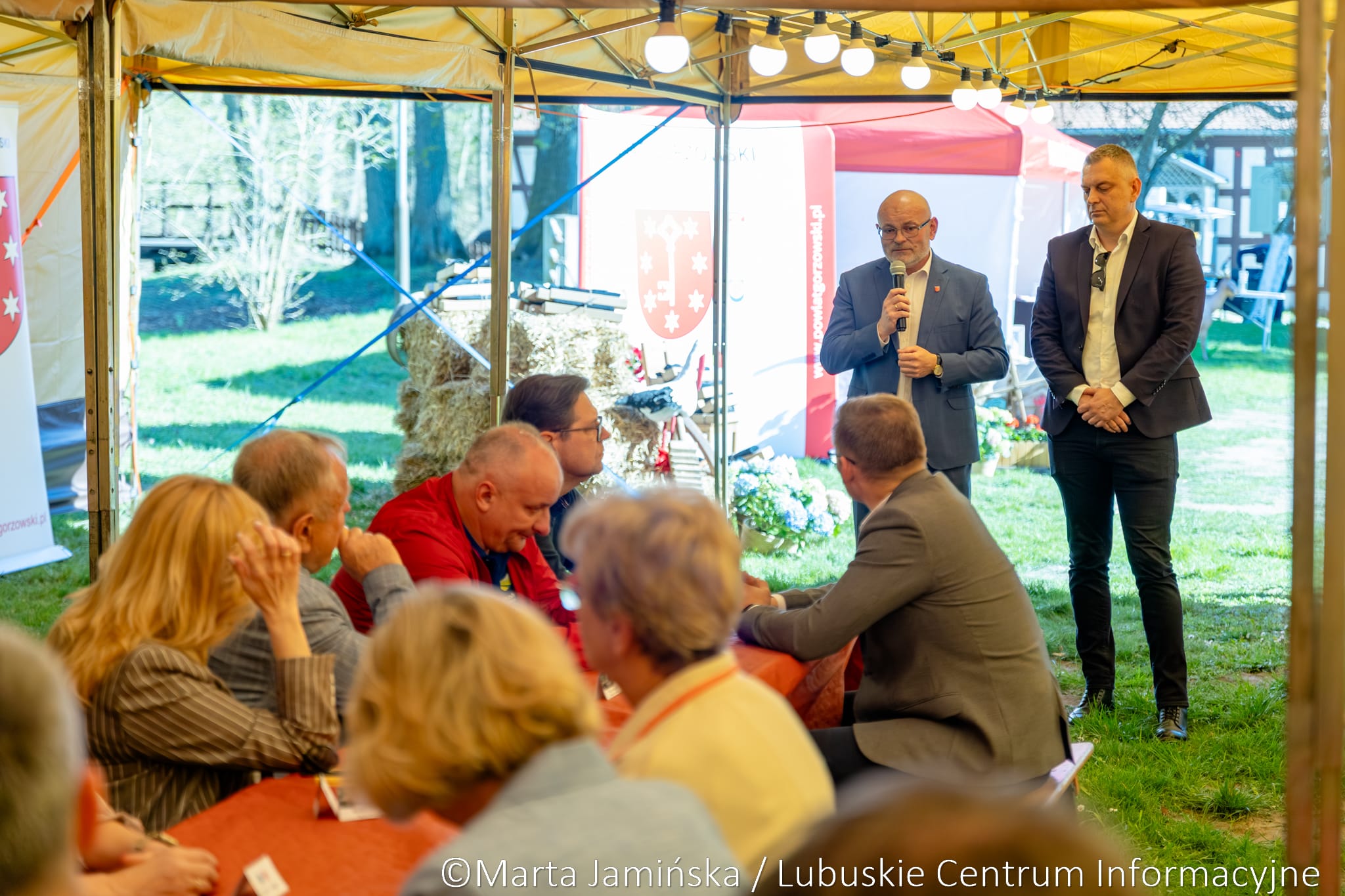 Man in a blue suit speaks into a microphone under a bright yellow canopy at an outdoor event, while another man stands nearby; audience sits at red‑cloth tables with banners in the background.