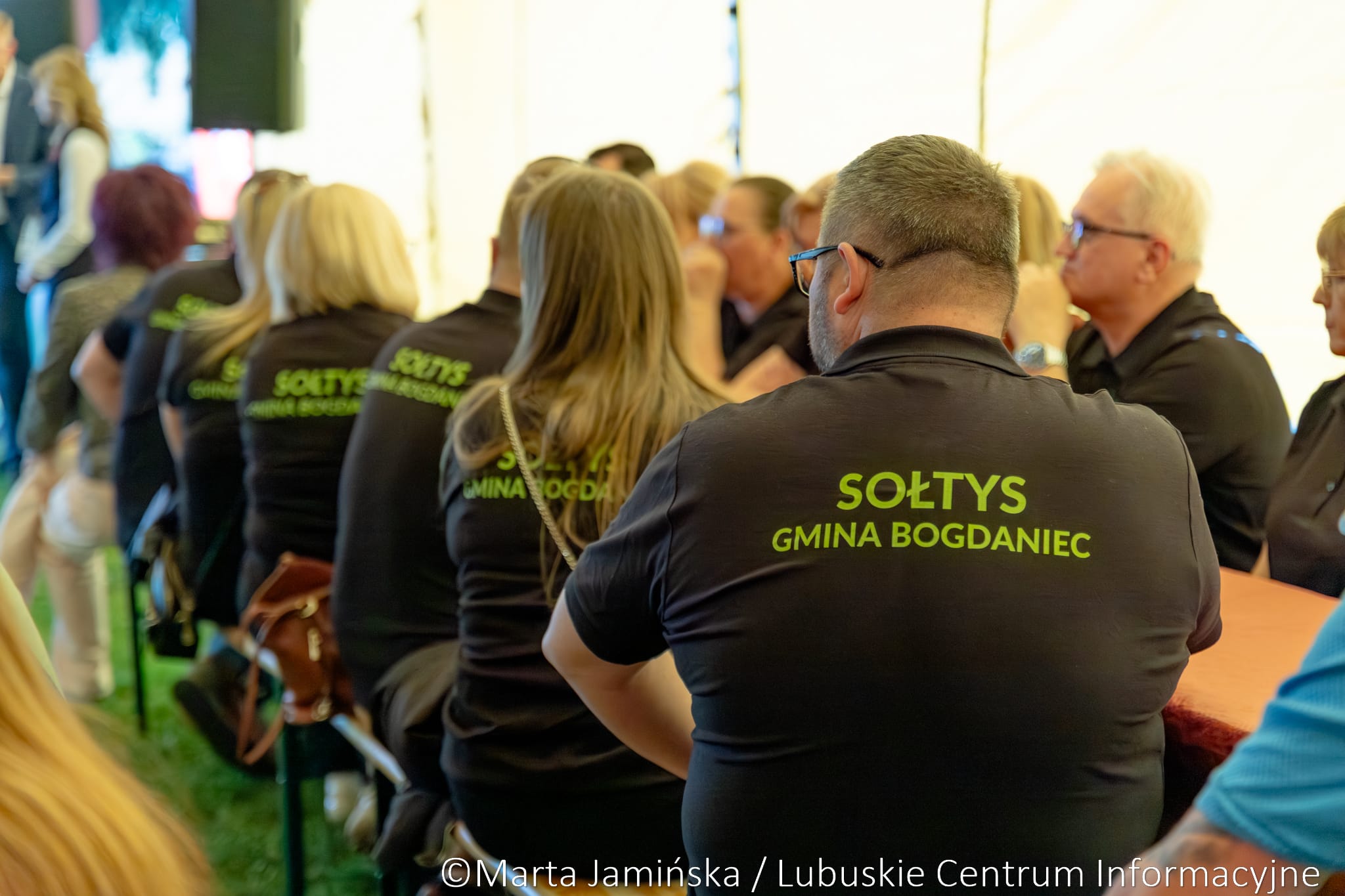Group of people wearing matching black shirts with lime text, attending a formal event or meeting outdoors/under a tent.