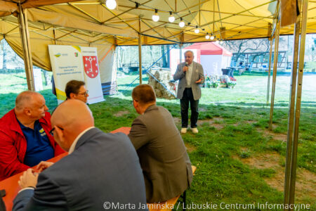 Man gives speech into a microphone under a yellow event tent while others sit at a table nearby. Bannered backdrop and outdoor scenery visible behind him.