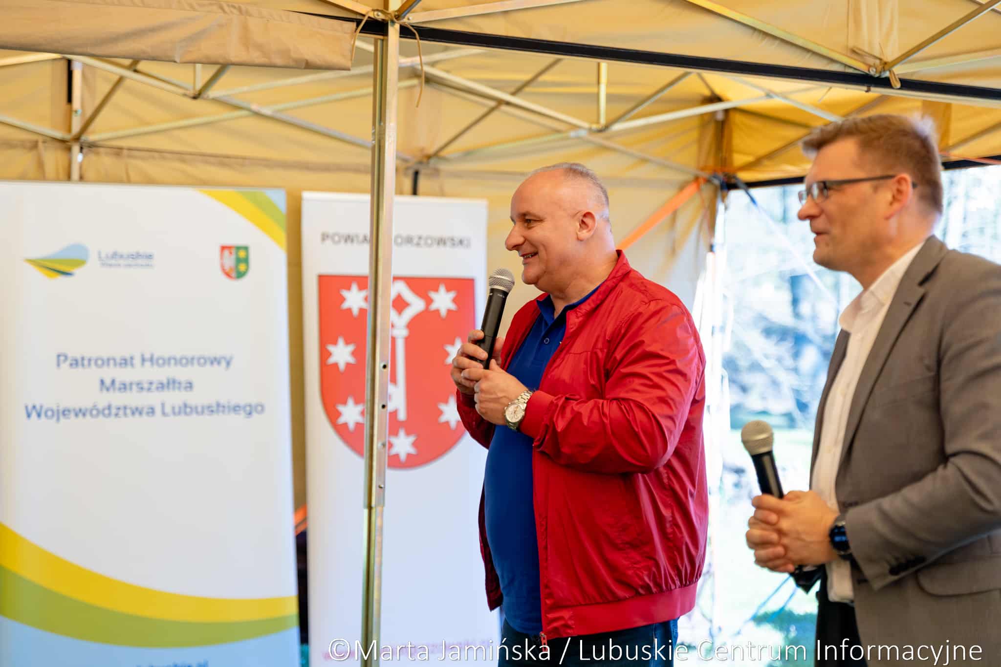 Two men stand on a stage under a beige canopy, each holding a microphone, with event banners in the background.