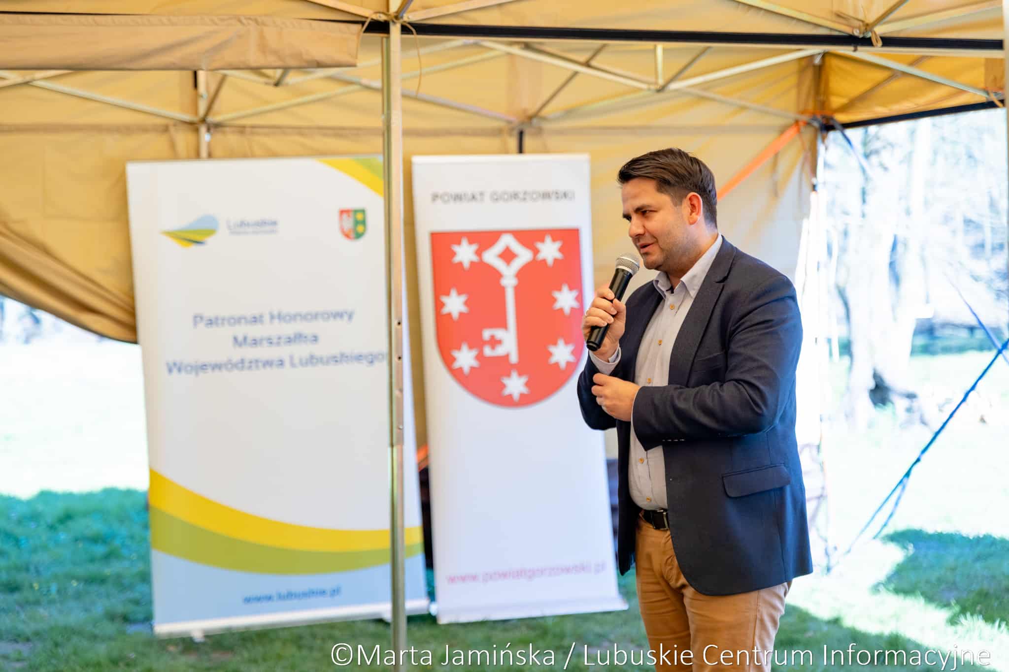 Man in a dark blazer speaks into a handheld microphone at an outdoor event under a beige tent, with banners in the background.