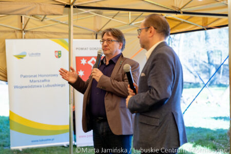 Two men in blazers speak into handheld microphones under a tent at an outdoor event, banners behind them