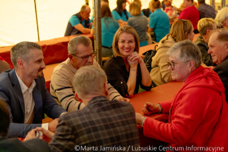 Group of adults seated at a long orange table under a tent, chatting and smiling; central woman wearing black smiles at the camera.