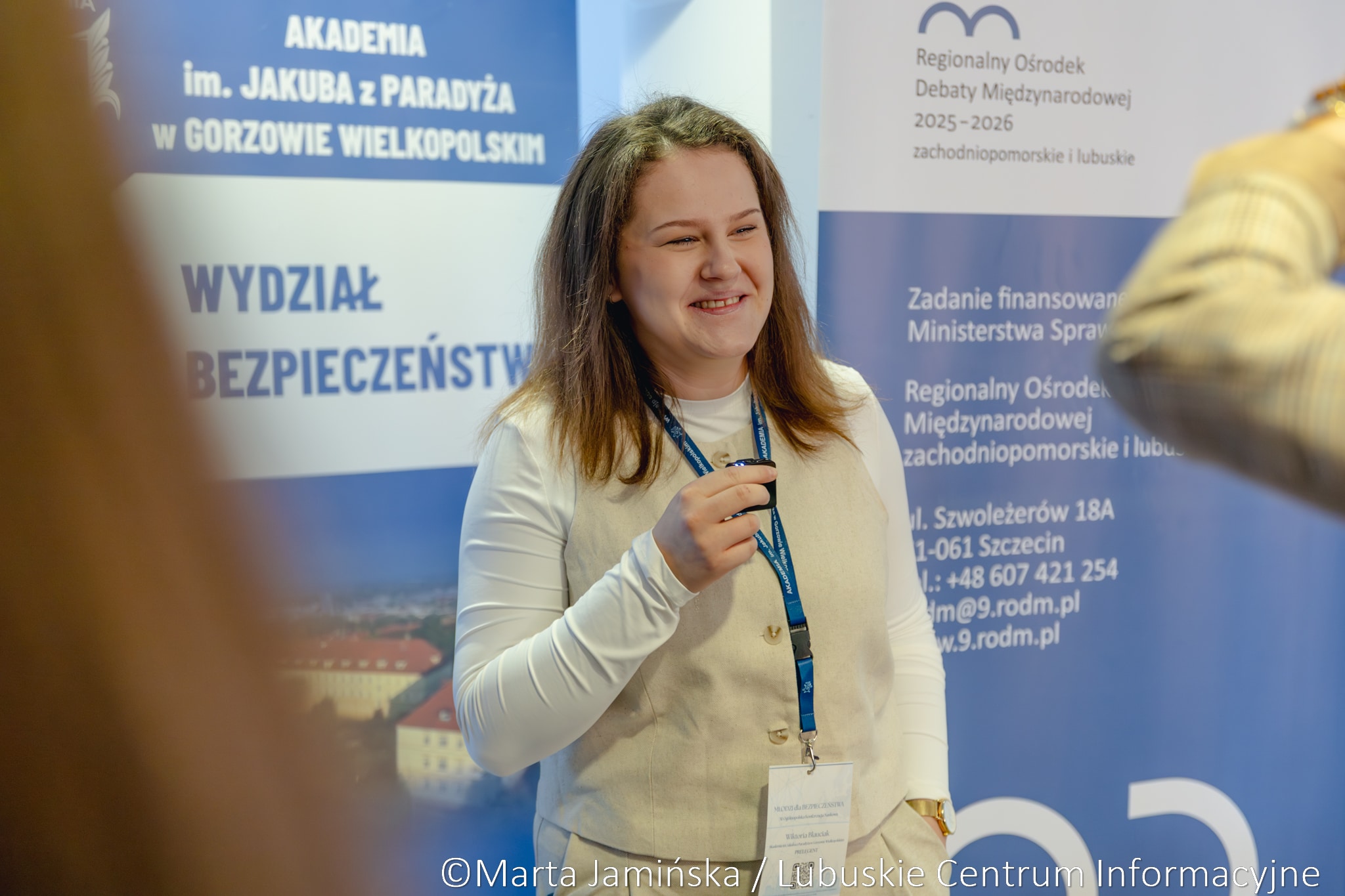 Young woman with a lanyard and name badge standing in front of blue informational banners at a conference, smiling.