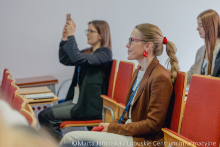 Audience members at a seminar: foreground woman in a brown blazer with red earrings and name badge, smiling and looking ahead.