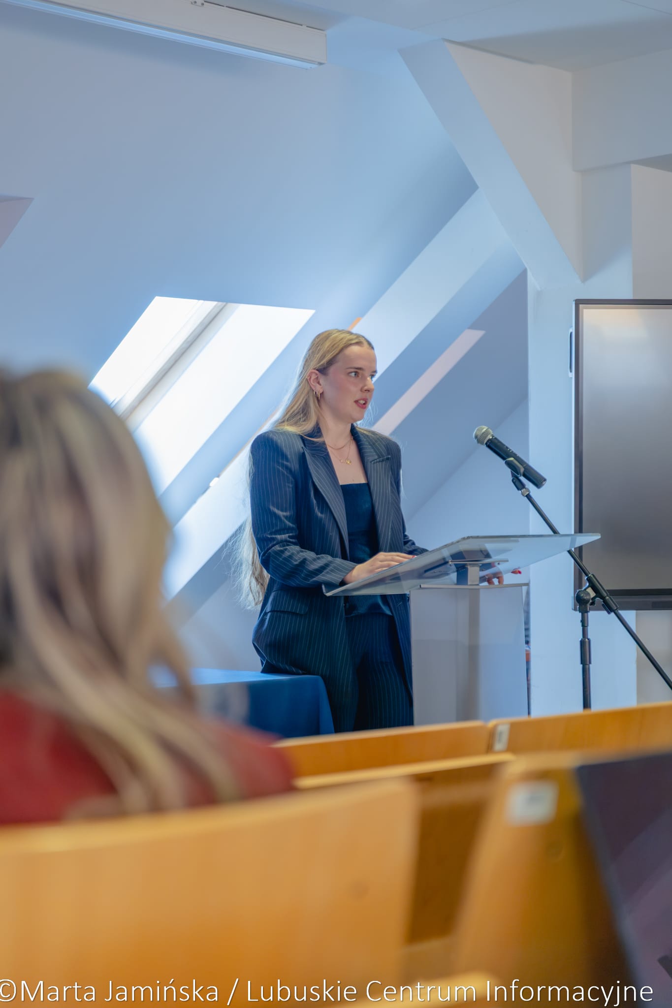 Woman in a blue pinstripe suit delivering a talk at a clear lectern with a microphone in a bright, modern conference room.
