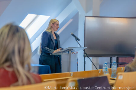 Woman in a blue pinstripe suit speaking at a podium with a microphone in a bright conference room.