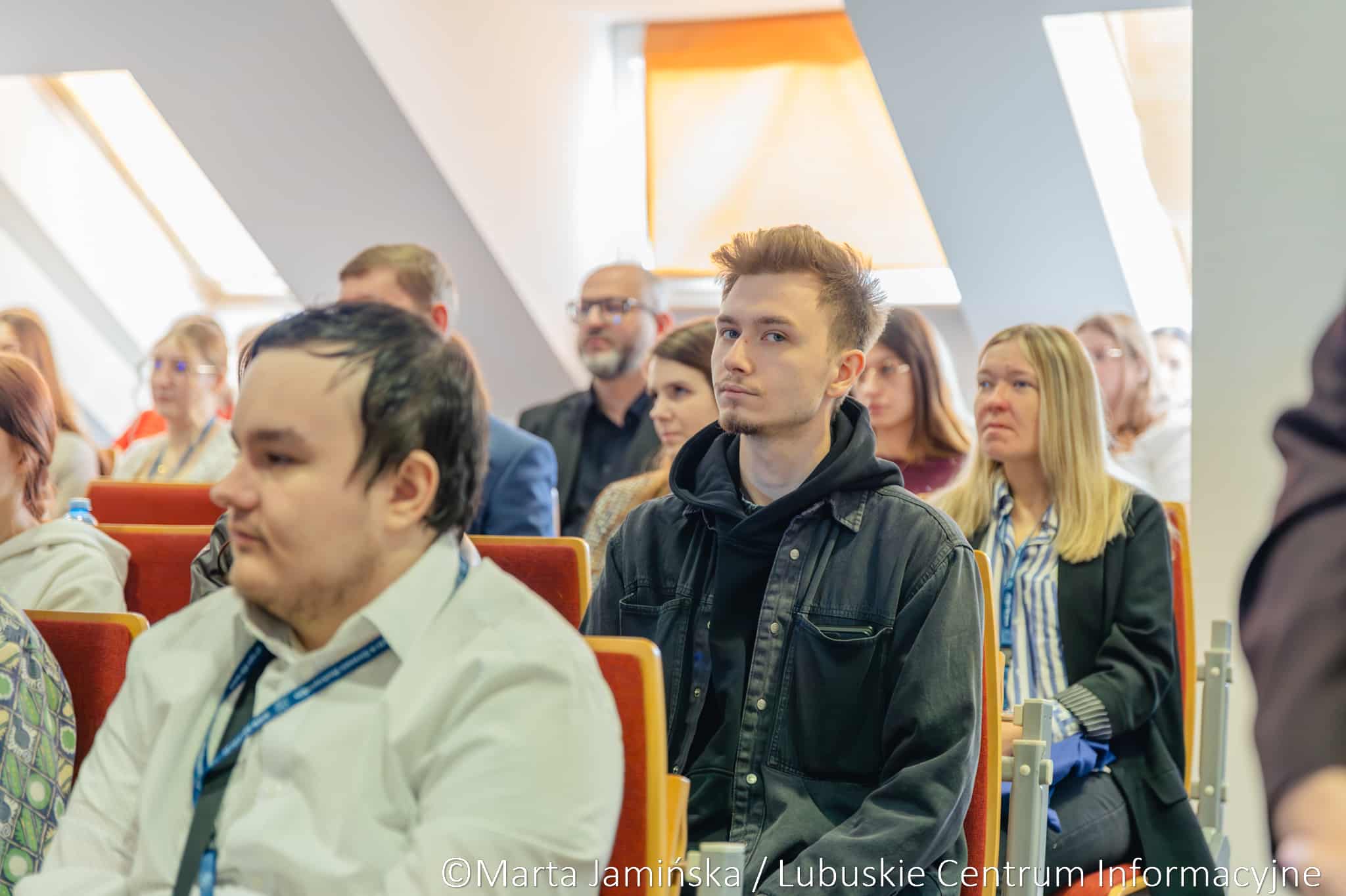 Audience members seated in orange chairs at a presentation, with a young man in a black denim jacket in the foreground.